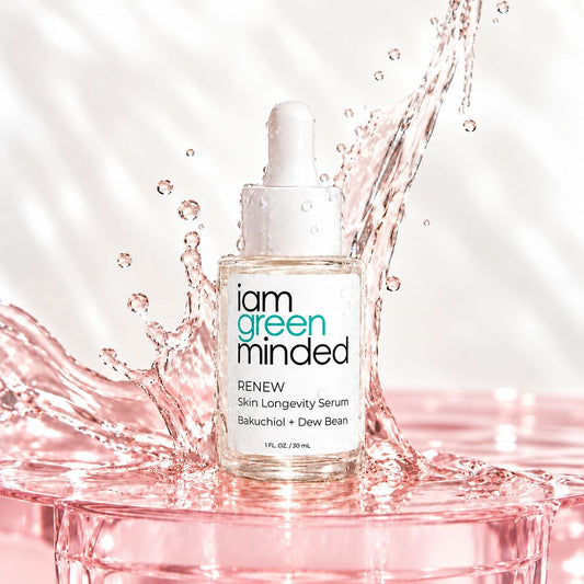 A clear glass dropper bottle of iam green minded RENEW Skin Longevity Serum stands on a pink translucent pedestal with a pink liquid splash behind it. The bottle has a white dropper cap and a white label with teal and black text.