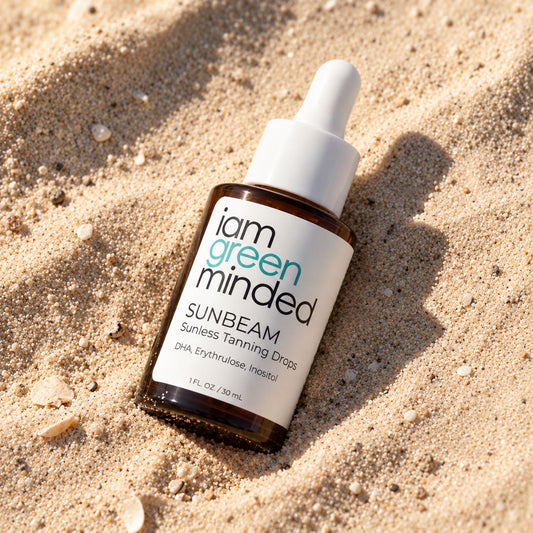 A bottle of iam green minded SUNBEAM Sunless Tanning Drops lies on sandy ground in bright sunlight, with the dropper cap pointing upward.