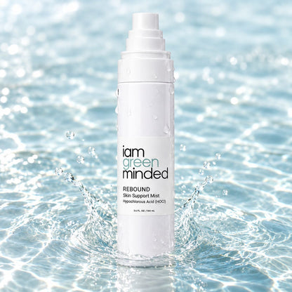 A white bottle of iam green minded REBOUND Skin Support Mist stands in splashing water, with droplets on the bottle and a bright blue water background.
