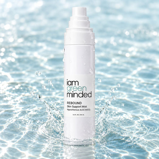 A white bottle of iam green minded REBOUND Skin Support Mist stands in splashing water, with droplets on the bottle and a bright blue water background.