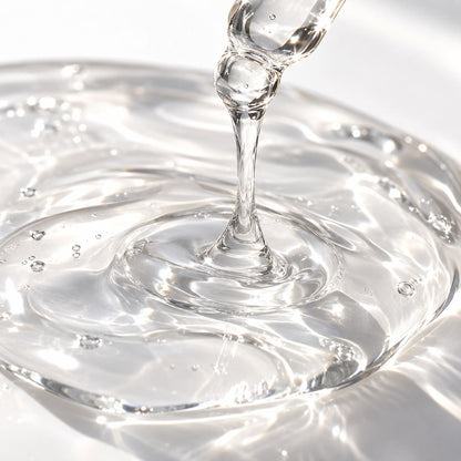 A close-up of a clear, viscous liquid being poured from a pipette, forming a ring of ripples and small bubbles on a white surface.