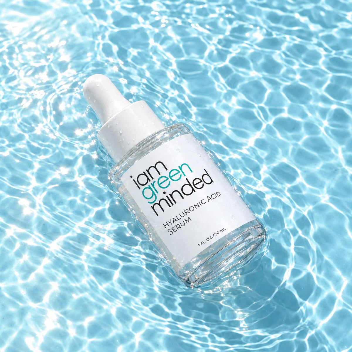 A clear glass dropper bottle of iam green minded Hyaluronic Acid Serum floating on clear blue water with bright sunlit ripples.