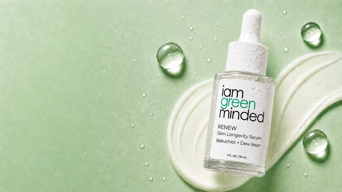 A clear glass dropper bottle of iam green minded Renew Skin Longevity Serum, placed on a white cream swirl with water droplets on a light green background.