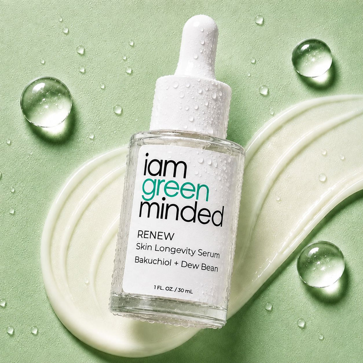 A bottle of iam green minded RENEW Skin Longevity Serum with a white dropper cap, set on a light green background with a cream-colored smear and water droplets.